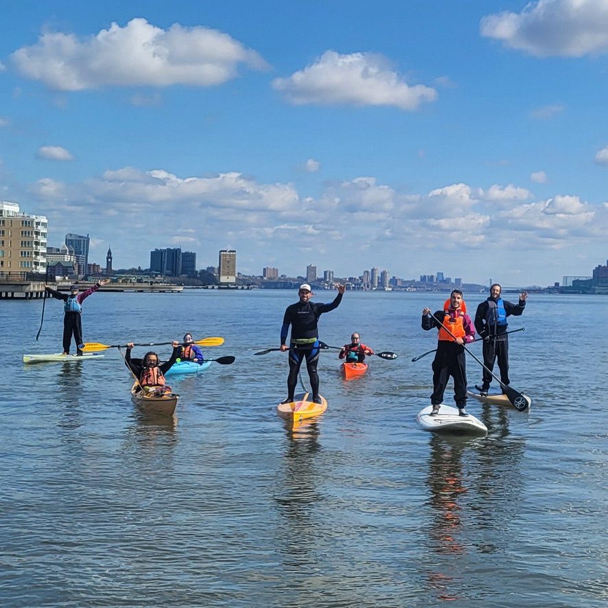 A group of paddlers on stand-up boards and in kayaks wave and raise their paddles on calm water, the waterfront behind them
