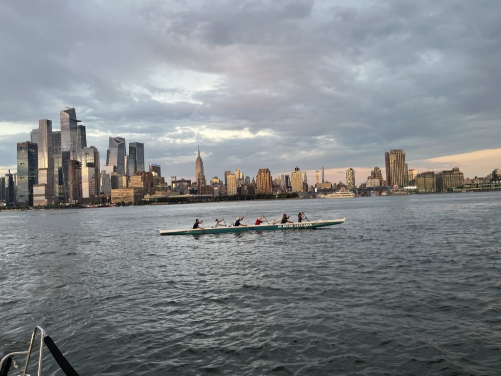 Six paddlers in an outrigger canoe on grey water in front of the Midtown skyline under a cloudy sky
