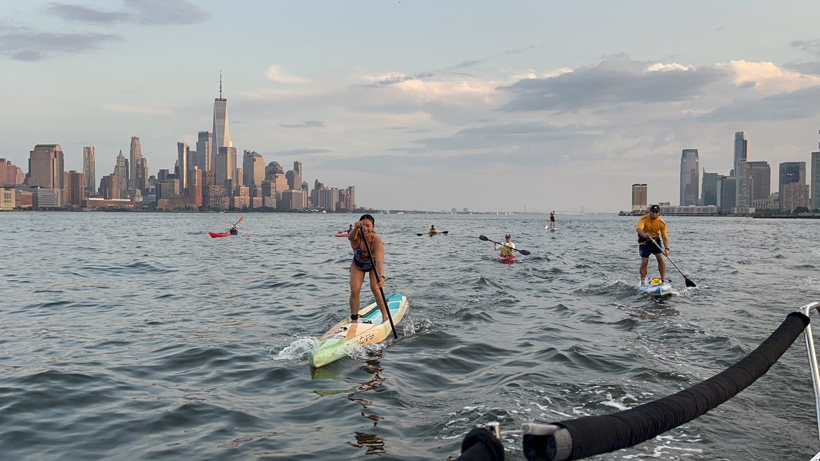 Two stand-up paddlers wearing belt life jackets ride wind chop on the Hudson, with kayakers and the Lower Manhattan skyline behind them