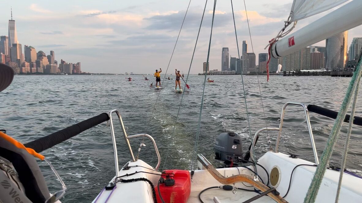 Looking back over the stern of a small sailboat at paddlers on stand-up boards and kayaks following in a long line down a choppy Hudson River at sunset, with the lower Manhattan skyline behind them