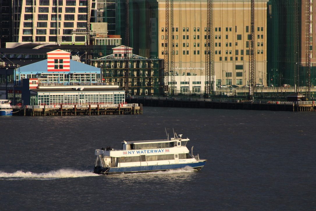 A NY Waterway ferry speeds past the Hoboken waterfront, a long white wake trailing behind it